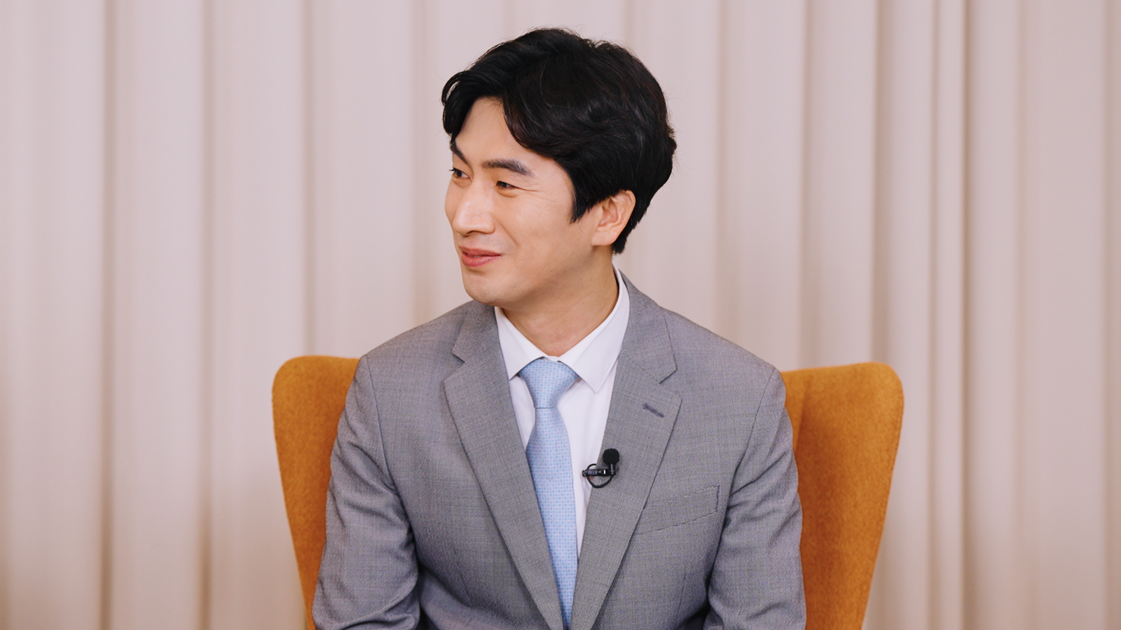 회색 정장에 넥타이를 매고 마이크를 착용한 남성이 인터뷰 중 미소 지으며 옆을 바라보고 있다.