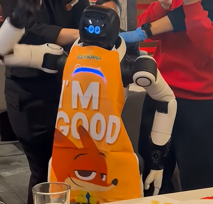 'I'M GOOD' 문구가 적힌 앞치마를 두른 휴머노이드 로봇이 여러 사람에게 둘러싸여 조작되는 모습.