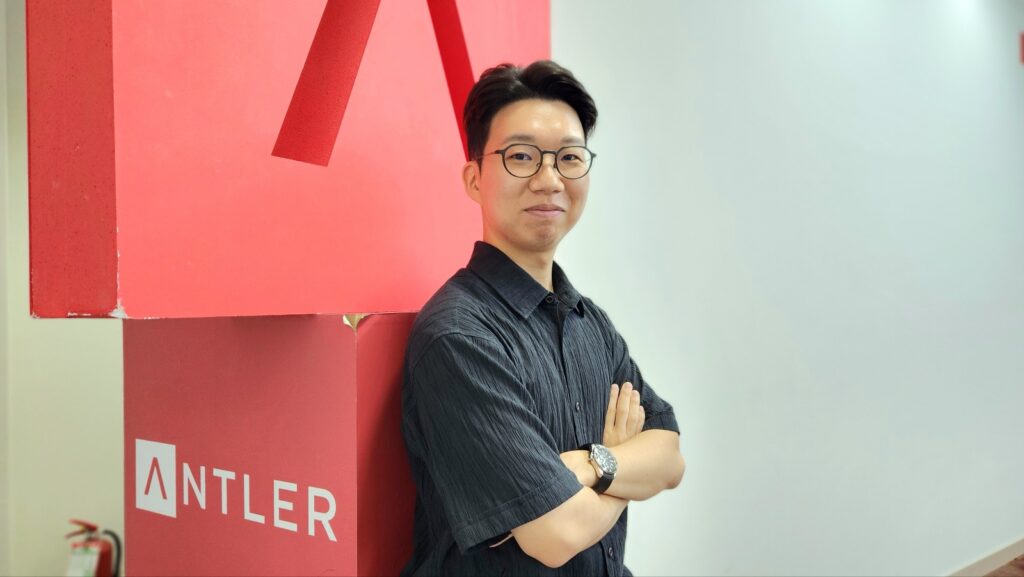 안경을 쓴 남성이 팔짱을 낀 채 앤틀러(ANTLER) 로고가 새겨진 붉은 조형물 앞에서 인터뷰 포즈를 취하고 있다.