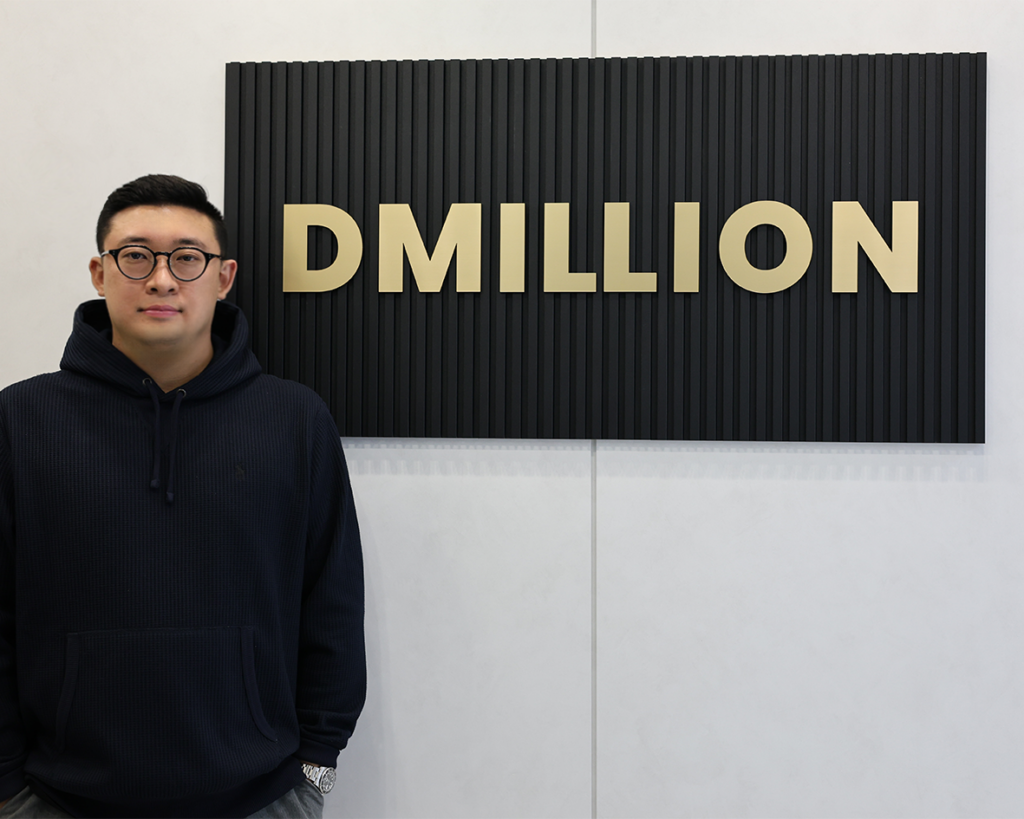 한요한 디밀리언 대표가 회사 로고 'DMILLION' 앞에서 서서 사진을 찍고 있다.