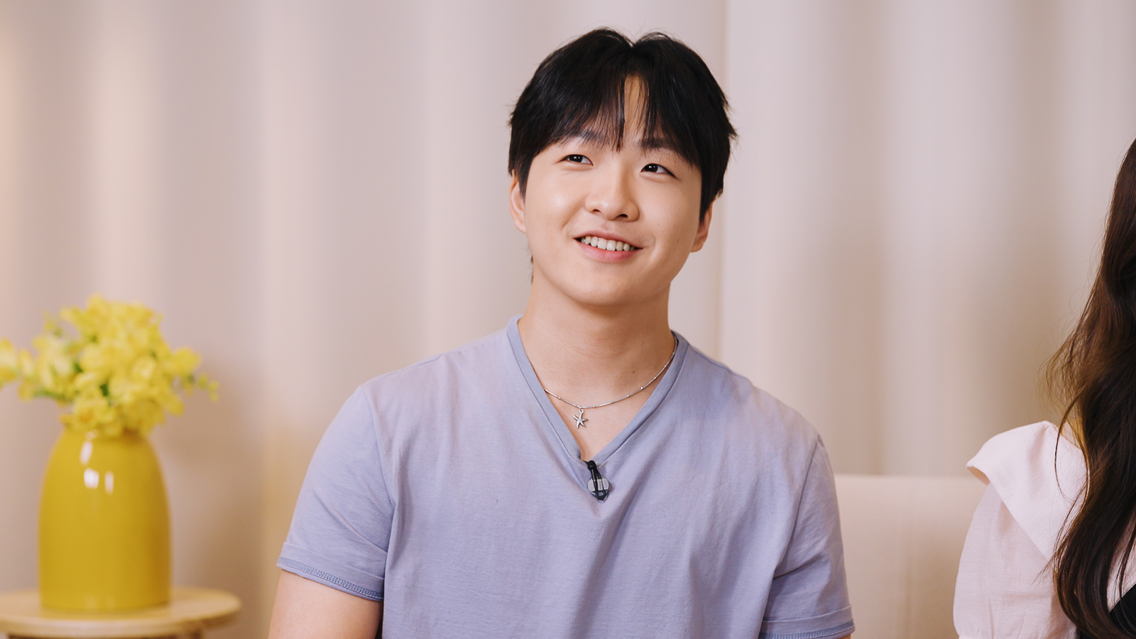 연보라색 티셔츠를 입은 남성이 인터뷰 중 밝게 웃고 있다.