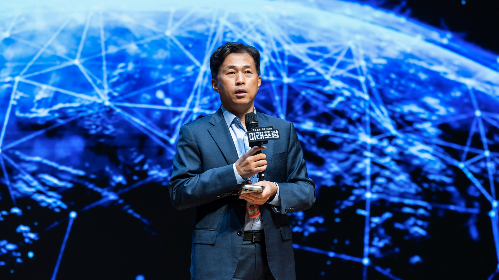 2026 SK하이닉스 미래포럼에서 관계자가 지구 네트워크 이미지를 배경으로 마이크를 들고 발표하고 있다.