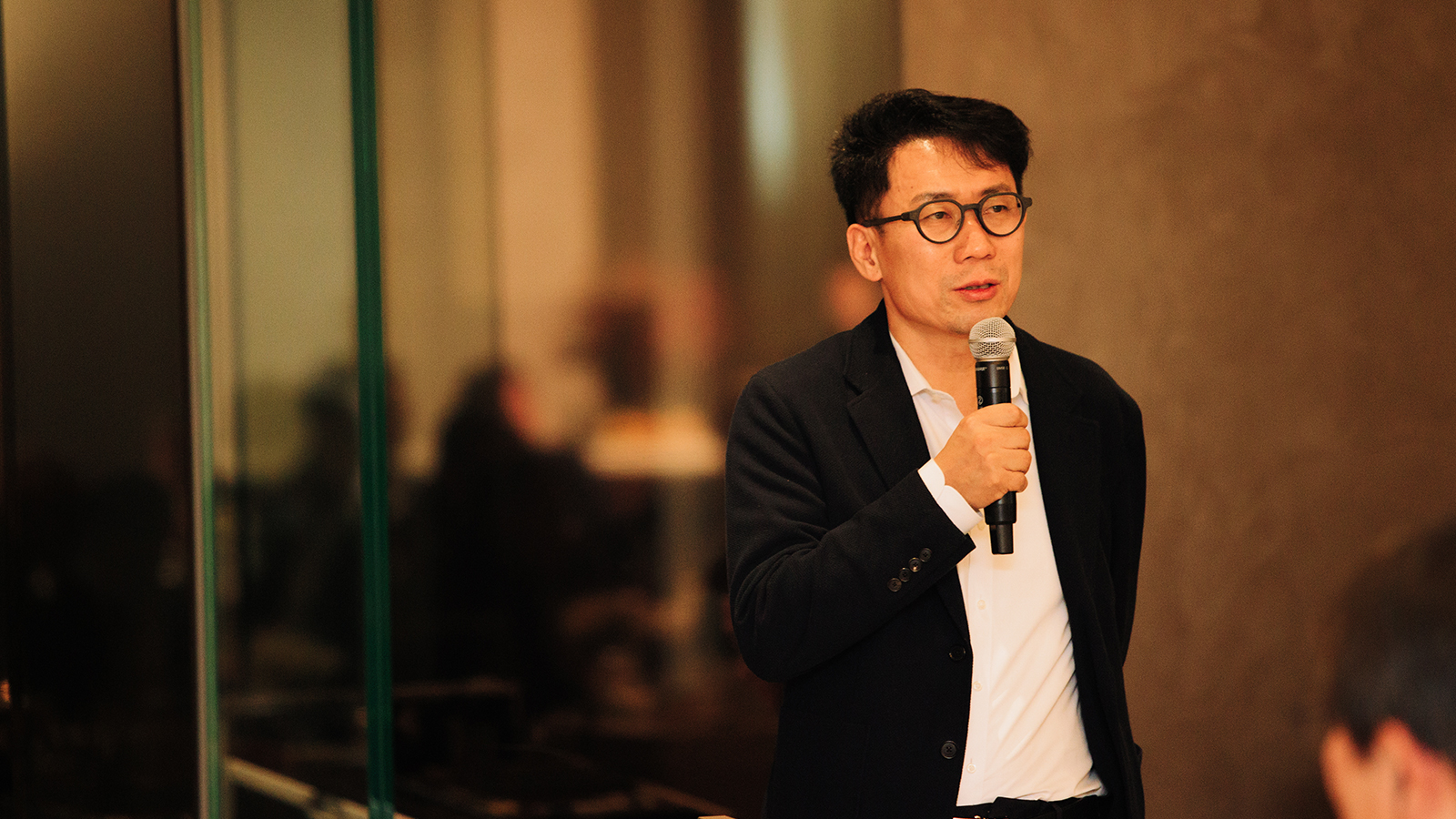 안경 쓴 남성이 마이크를 들고 행사에서 발언하고 있다.