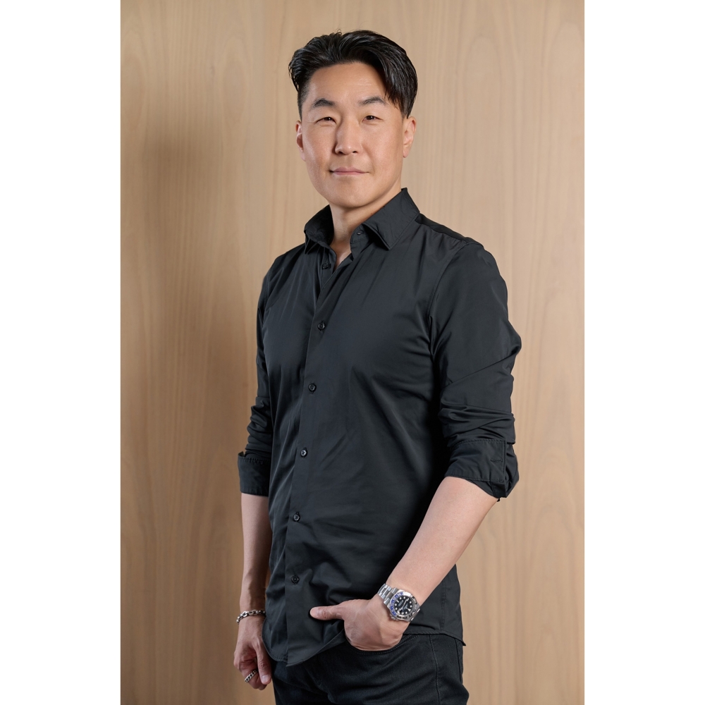 검은색 셔츠를 입은 남성이 목재 배경 앞에서 정면을 바라보며 서 있는 인물 사진.