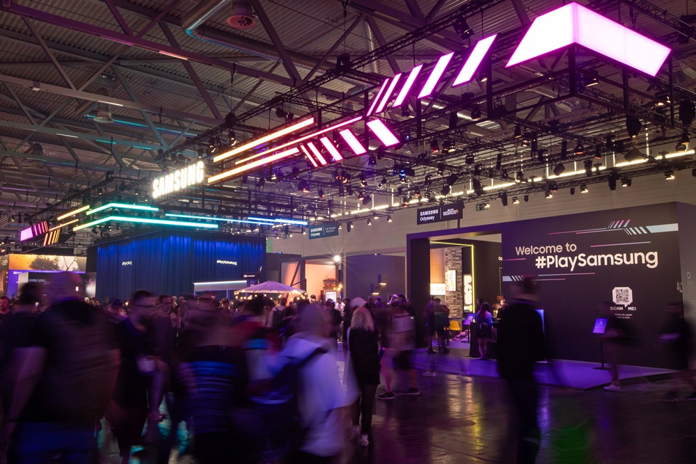 네온 조명이 화려한 삼성 부스에 관람객들이 모여있고, 'Welcome to #PlaySamsung' 문구가 보인다.
