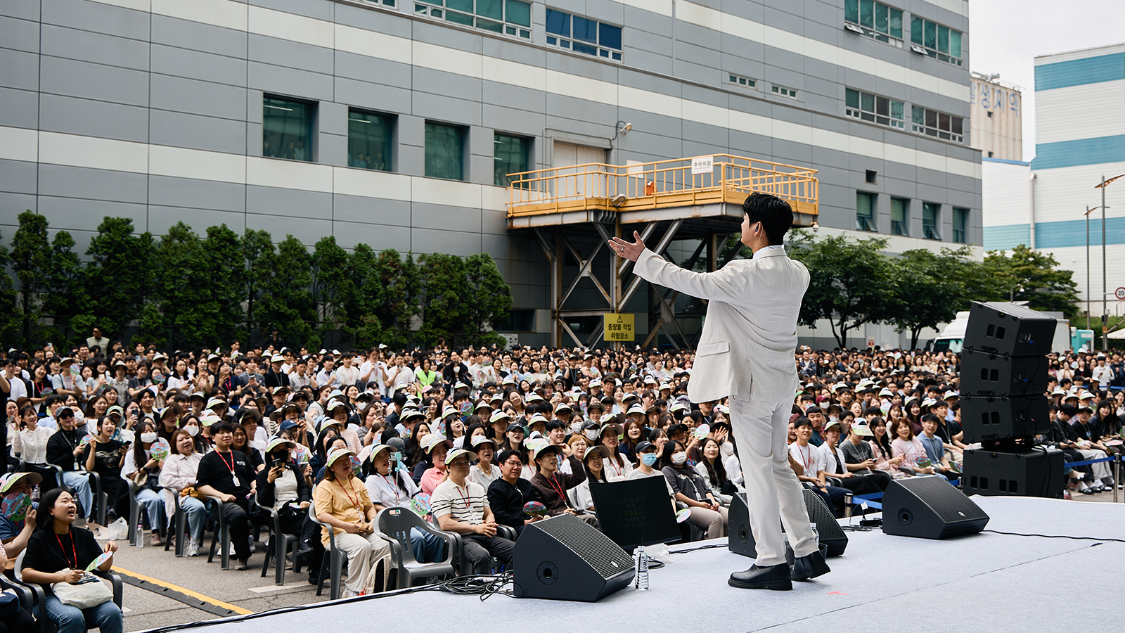 흰색 정장을 입은 남성이 야외 무대에서 팔을 들어 인사하며, 수많은 관객이 모자를 쓰고 앉아 공연을 지켜보고 있다.