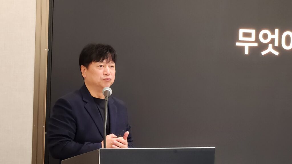 남색 재킷 차림의 발표자가 어두운 배경 무대에서 마이크를 잡고 강연하는 모습.
