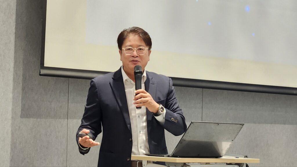 안경 쓴 남성이 마이크를 들고 스크린 앞에서 스타트업 M&A 관련 강연을 하고 있다.