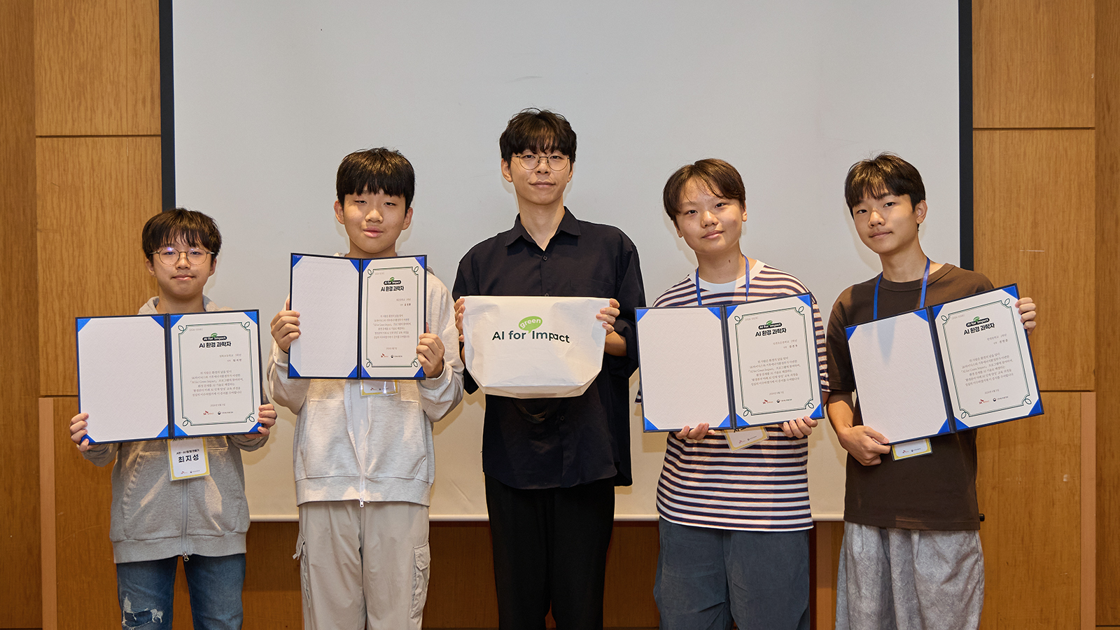 'AI 환경 과학자' 인증서를 든 학생 4명과 관계자가 'AI for Impact' 에코백을 들고 기념 촬영을 하고 있다.