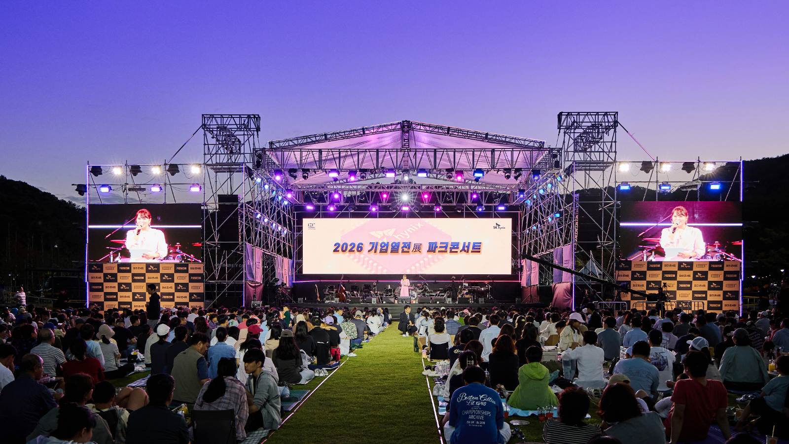 '2026 기업열전展 파크콘서트' 무대에서 가수가 공연하고 잔디밭에 앉은 관람객들이 관람하는 모습.