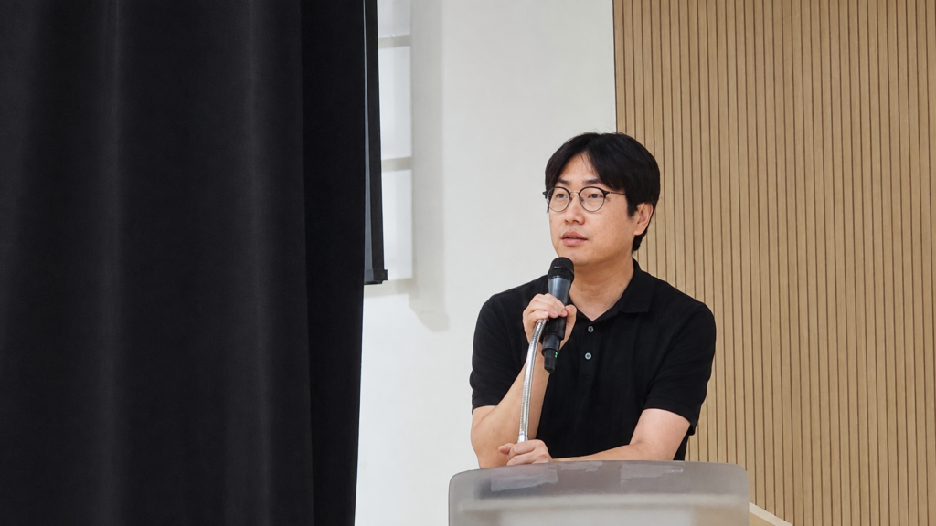 안경을 쓴 남성이 검은색 옷을 입고 마이크를 들고 강단에서 발언하고 있다.
