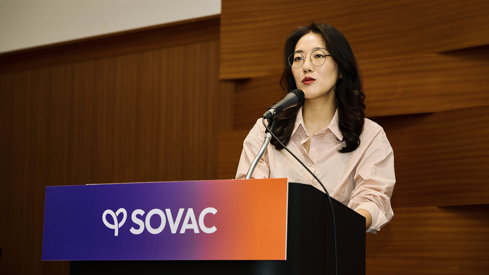 SOVAC 연단에서 안경을 쓴 여성 발표자가 마이크 앞에서 발언하고 있다.