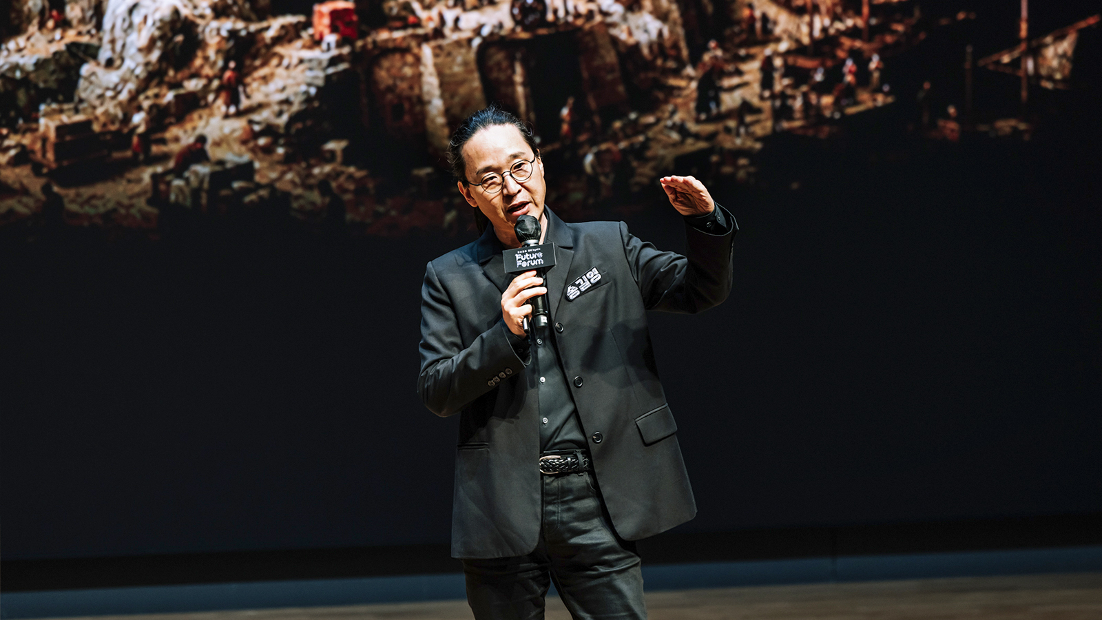 연사가 마이크를 들고 손짓하며 발표하는 모습, 뒤편 화면에는 대규모 인파 장면이 펼쳐져 있다.