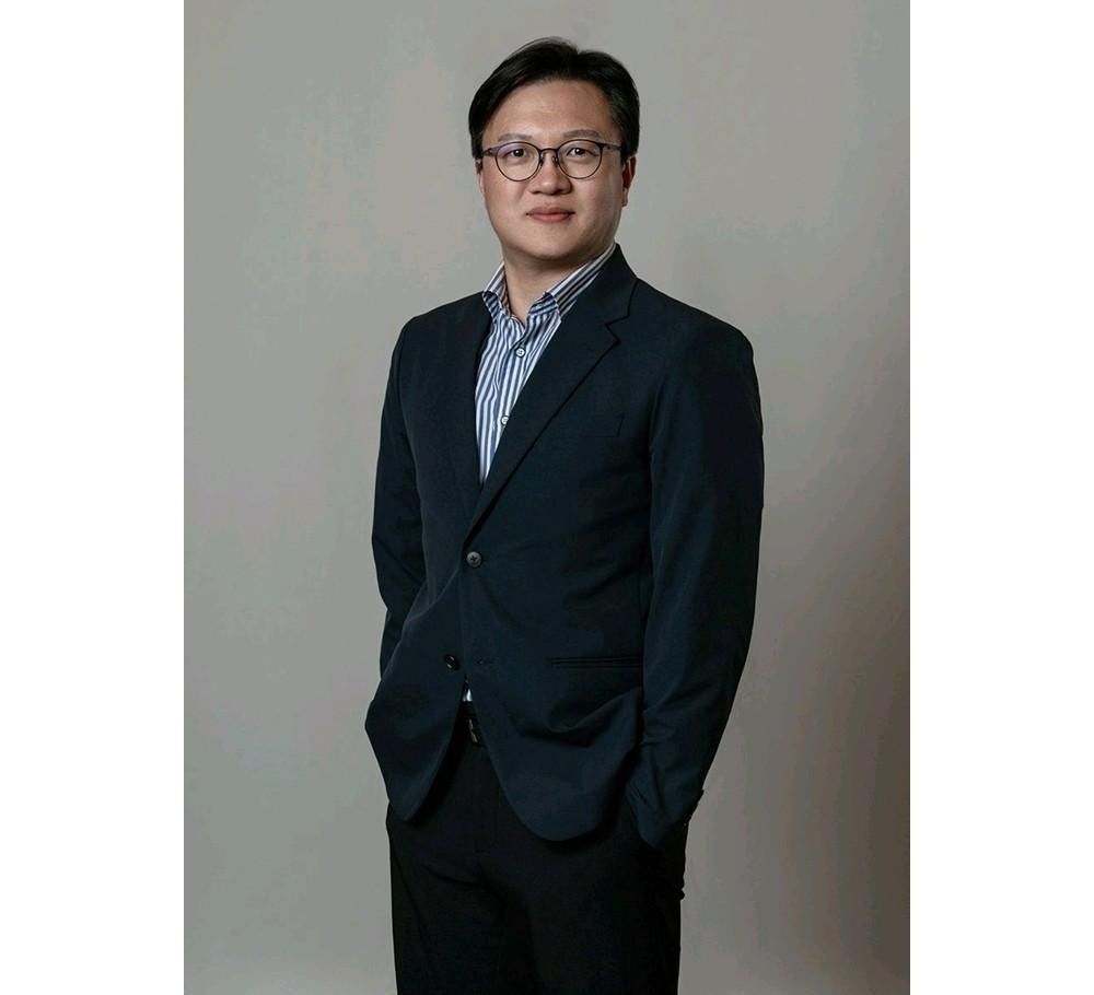 안경을 쓴 남성이 짙은 색 재킷 차림으로 정면을 바라보며 서 있는 인물 사진.