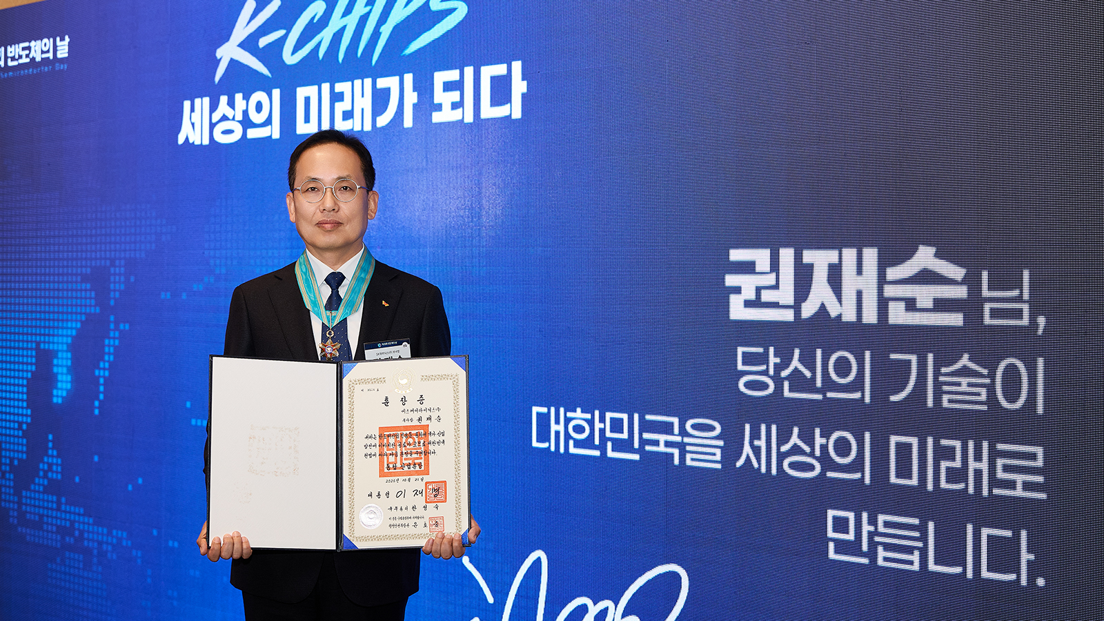 반도체의 날 행사에서 훈장을 목에 걸고 훈장증을 든 권재순 씨가 'K-CHIPS' 배경 앞에서 기념 촬영을 하고 있다.