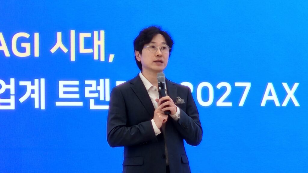 안경 쓴 남성이 'AGI 시대, 업계 트렌드 2027 AX'가 적힌 파란 배경 앞에서 마이크를 들고 발표하고 있다.